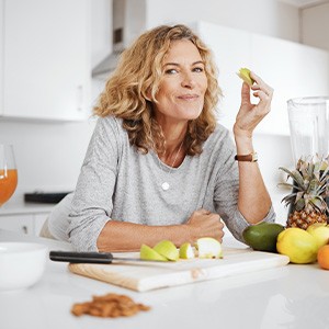 Woman in gray shirt leaning on white kitchen counter eating slice of green apple