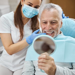 Man with white hair smiling at handheld mirror as dentist shows him his teeth