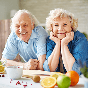 Older man and woman in blue shirts at kitchen counter with several fruits and a cutting board