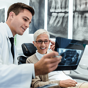 Woman with white hair in dental chair touching glasses looking at X-rays with dentist